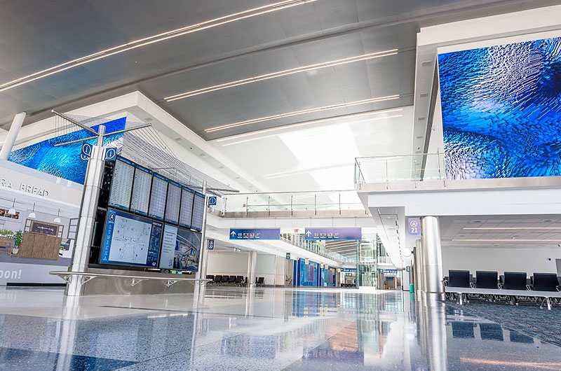A bright modern airport terminal featuring large overhead Nanolumens LED video walls displaying blue abstract imagery